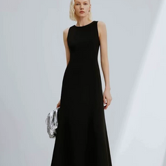 A woman modeling a sleeveless black midi dress with a high neckline, holding a small silver patterned clutch against a light gray studio background.