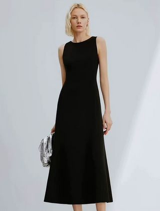 A woman modeling a sleeveless black midi dress with a high neckline, holding a small silver patterned clutch against a light gray studio background.