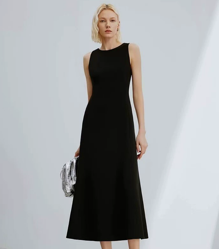 A woman modeling a sleeveless black midi dress with a high neckline, holding a small silver patterned clutch against a light gray studio background.