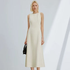 A woman modeling a sleeveless, high-neck midi dress in a warm off-white or cream colour, holding a small black handbag against a minimalist light gray background with soft diagonal shadows.