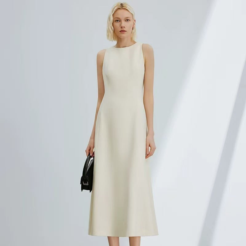 A woman modeling a sleeveless, high-neck midi dress in a warm off-white or cream colour, holding a small black handbag against a minimalist light gray background with soft diagonal shadows.