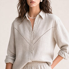 A woman wearing a light beige linen button-up shirt with delicate black dotted stitching details across the chest, styled with a pearl necklace and white earrings against a soft beige background.