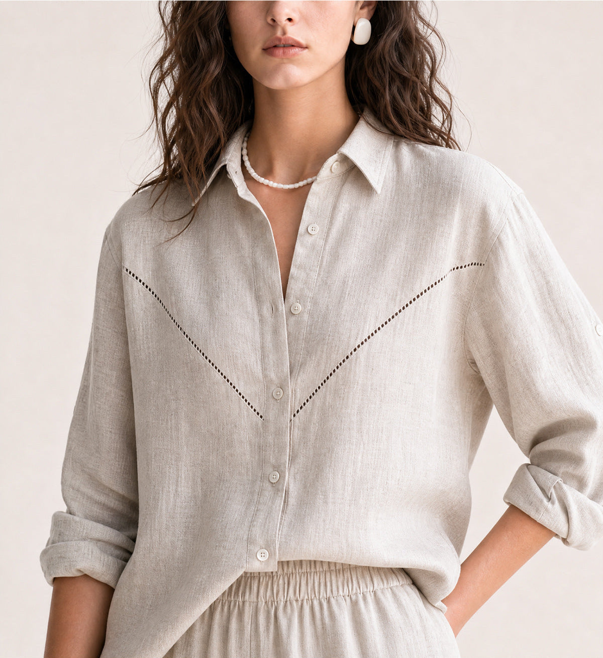 A woman wearing a light beige linen button-up shirt with delicate black dotted stitching details across the chest, styled with a pearl necklace and white earrings against a soft beige background.